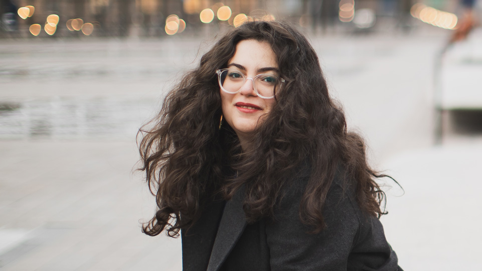 İlayda Deniz Oğuz - A women with dark brown curly hair and glasses, wearing a black coat, smiling at the camera. İlayda Deniz Oğuz - A women with dark brown curly hair and glasses, wearing a black coat, smiling at the camera.